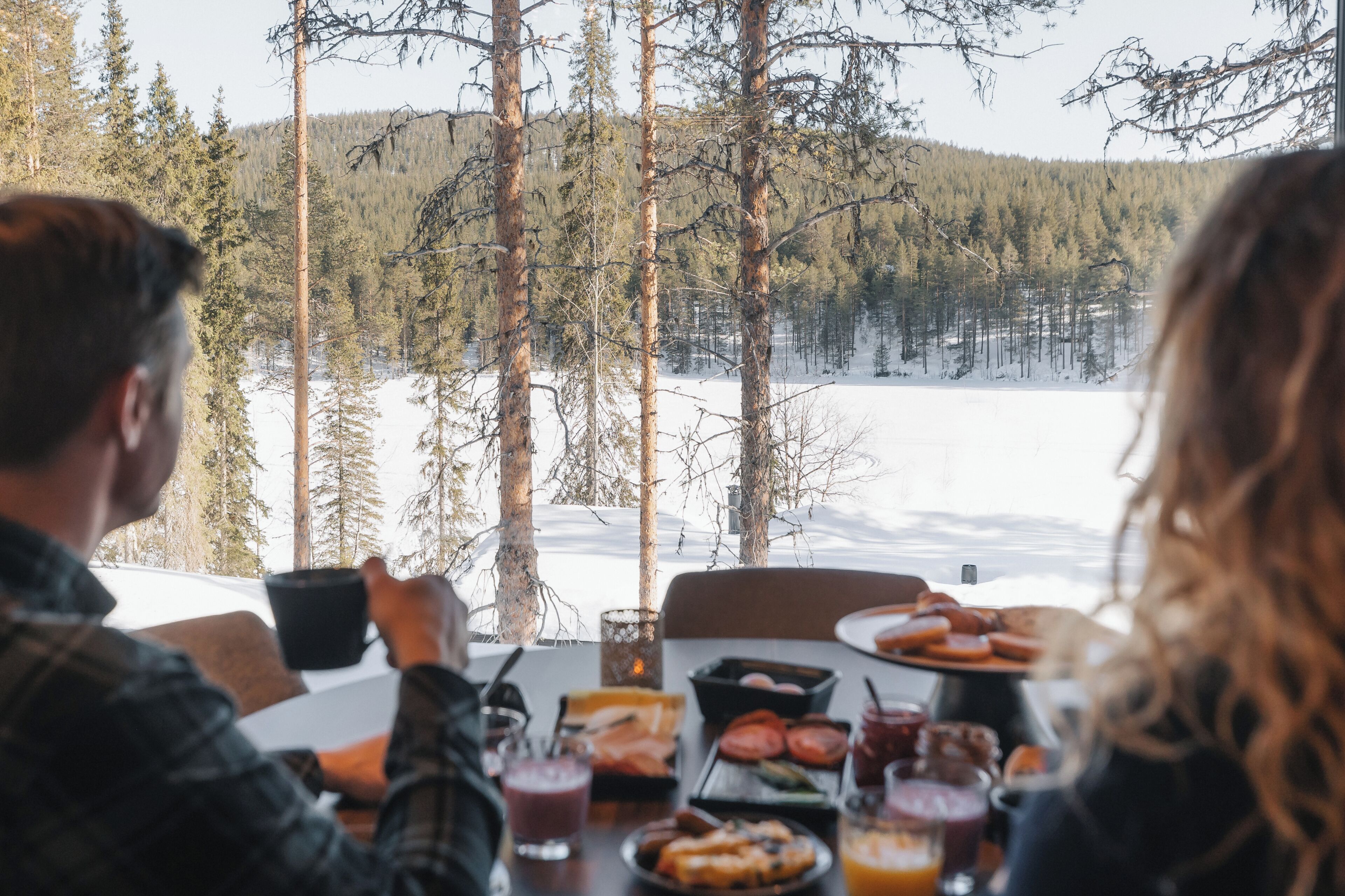 Ein Esstisch mit Speisen und Getränken, mit Blick auf eine verschneite Landschaft mit Bäumen.