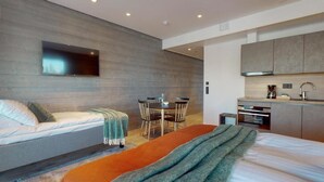 A modern hotel room with a bed, a small dining table, a sofa, a TV mounted on the wall, and a kitchenette.