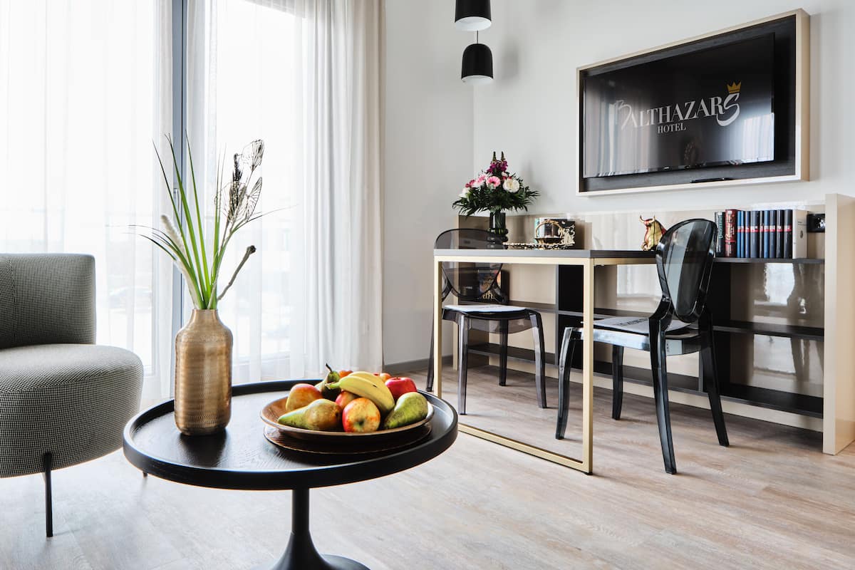 Een moderne hotelkamer met een zithoek, een groot raam met gordijnen, een flatscreen televisie aan de muur, en een vaas met bloemen op een tafel.