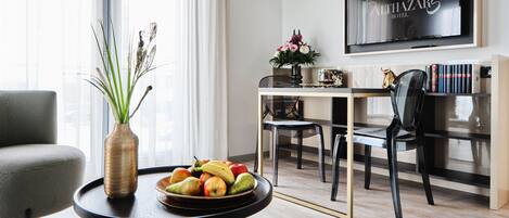 A modern hotel room with a dining area, a large window with curtains, a flat-screen TV on the wall, and a vase with flowers on a table.