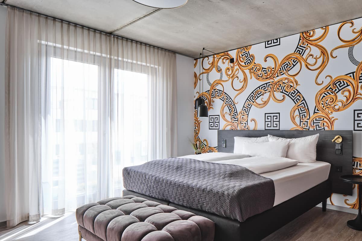 Een moderne slaapkamer met een groot bed, een gestoffeerde poef en een decoratieve muur met een geometrisch patroon.