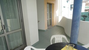 Ein kleiner Balkon mit gefliestem Boden, weißen Plastikstühlen, einem schwarzen Tisch und einer gelben Tischmatte mit zwei Glaskannnen.