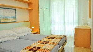 Un dormitorio con cama, mesita de noche, escritorio y ventana con cortinas traslúcidas.