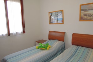 A room with two beds, a small bedside table, a window with curtains, and two framed pictures on the wall. - Cozy flat with swimming pools-Beahost (Bibione)