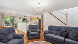 A modern living room with grey sofas, a chandelier, and a kitchen in the background. - Safety Beach Cottage (Safety Beach)