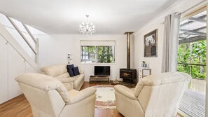 A living room with a sofa set, a TV, a fireplace, and a large window with a view of greenery. - Safety Beach Cottage (Safety Beach)