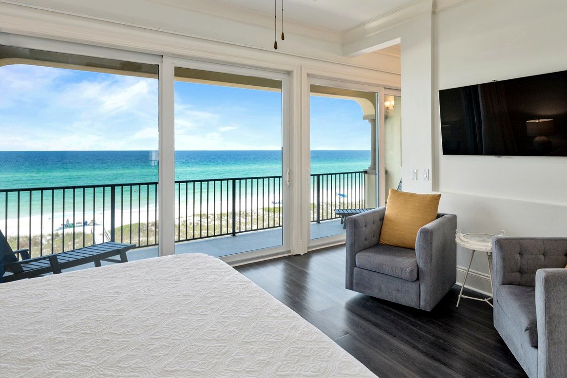Habitación de hotel con una cama grande, un sillón gris, una mesita y vistas a la playa y al océano.