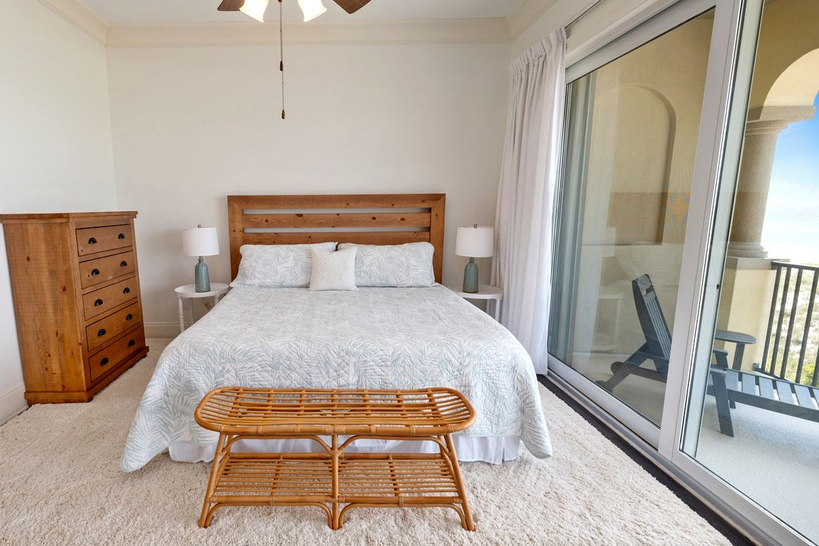 Une chambre avec un grand lit, des lampes de chevet, une commode en bois, un banc en osier et un balcon avec une chaise.
