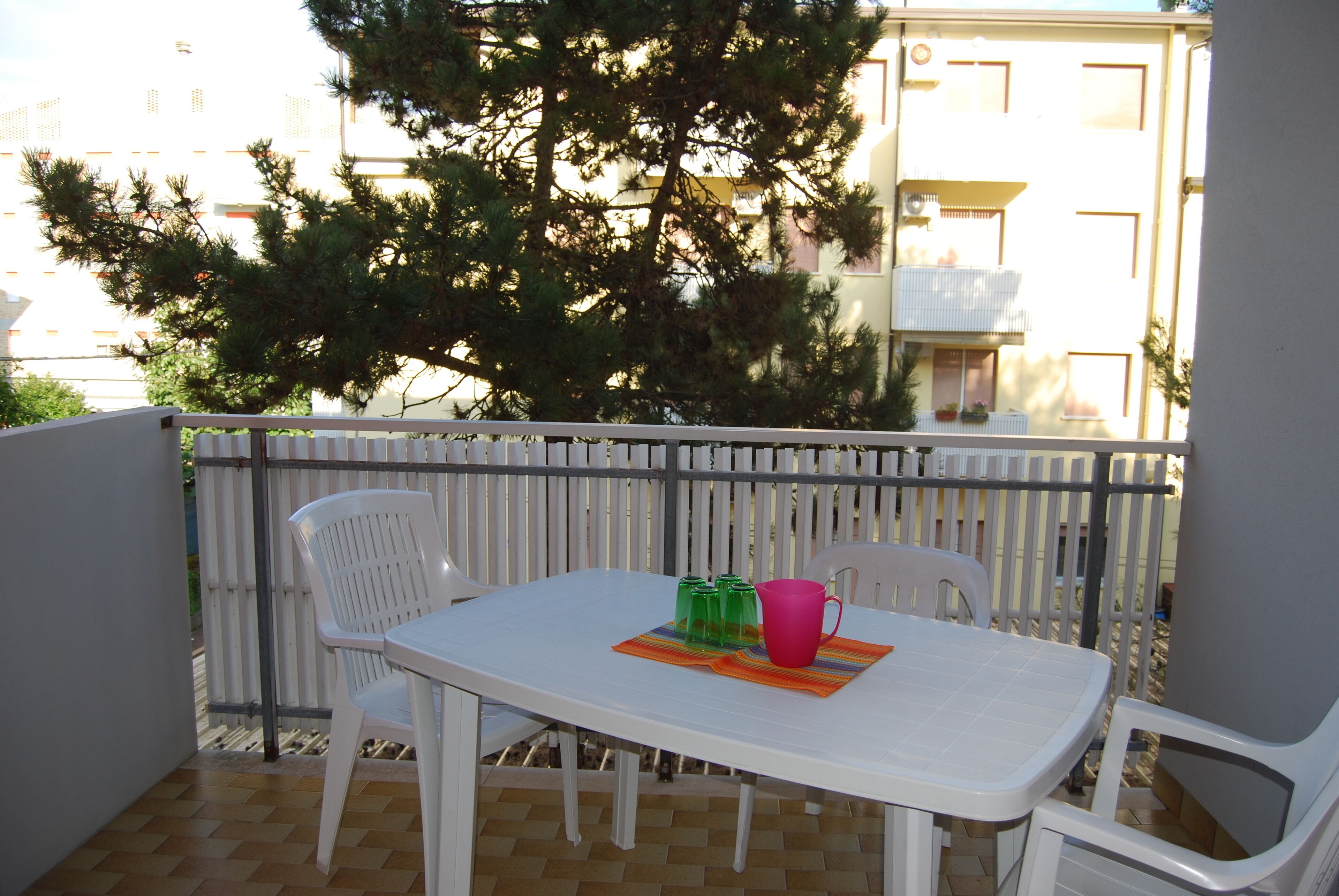 Un balcón con una mesa y sillas blancas, barandilla de metal y un árbol grande al fondo.