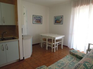 A compact kitchen with a sink, a refrigerator, a dining table with chairs, and two framed pictures on the wall.