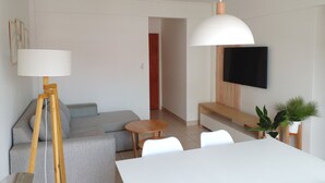 A modern living room with a grey sofa, a wooden coffee table, a white dining table, and a TV mounted on the wall.