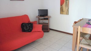 Una stanza con un divano rosso, un mobile TV in legno con televisore a schermo piatto, un tavolo da pranzo con sedie e due quadri floreali alle pareti.