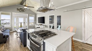 A modern kitchen with a central island, a ceiling fan, and a large window overlooking a deck. - Cozy Bay House w/ Heated Spool + Pier (San Leon)
