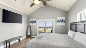 A bedroom with a large bed, a ceiling fan, a bench, and a view of the outdoors. - Cozy Bay House w/ Heated Spool + Pier (San Leon)