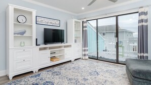 Un salon moderne doté d’un téléviseur à écran plat, de meubles blancs, d’un ventilateur de plafond et d’une grande porte-fenêtre coulissante donnant sur un balcon.