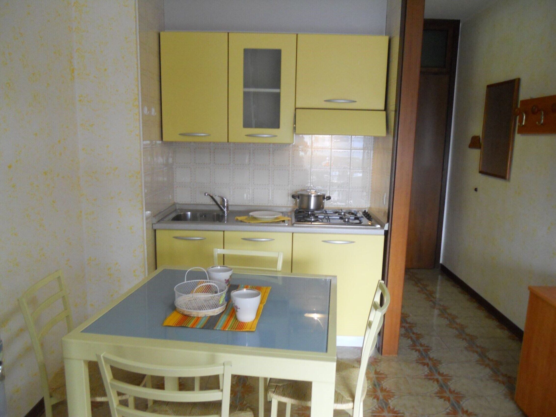 Une petite cuisine avec des placards jaunes, un évier et une cuisinière. On y trouve une table avec des chaises et un tapis.