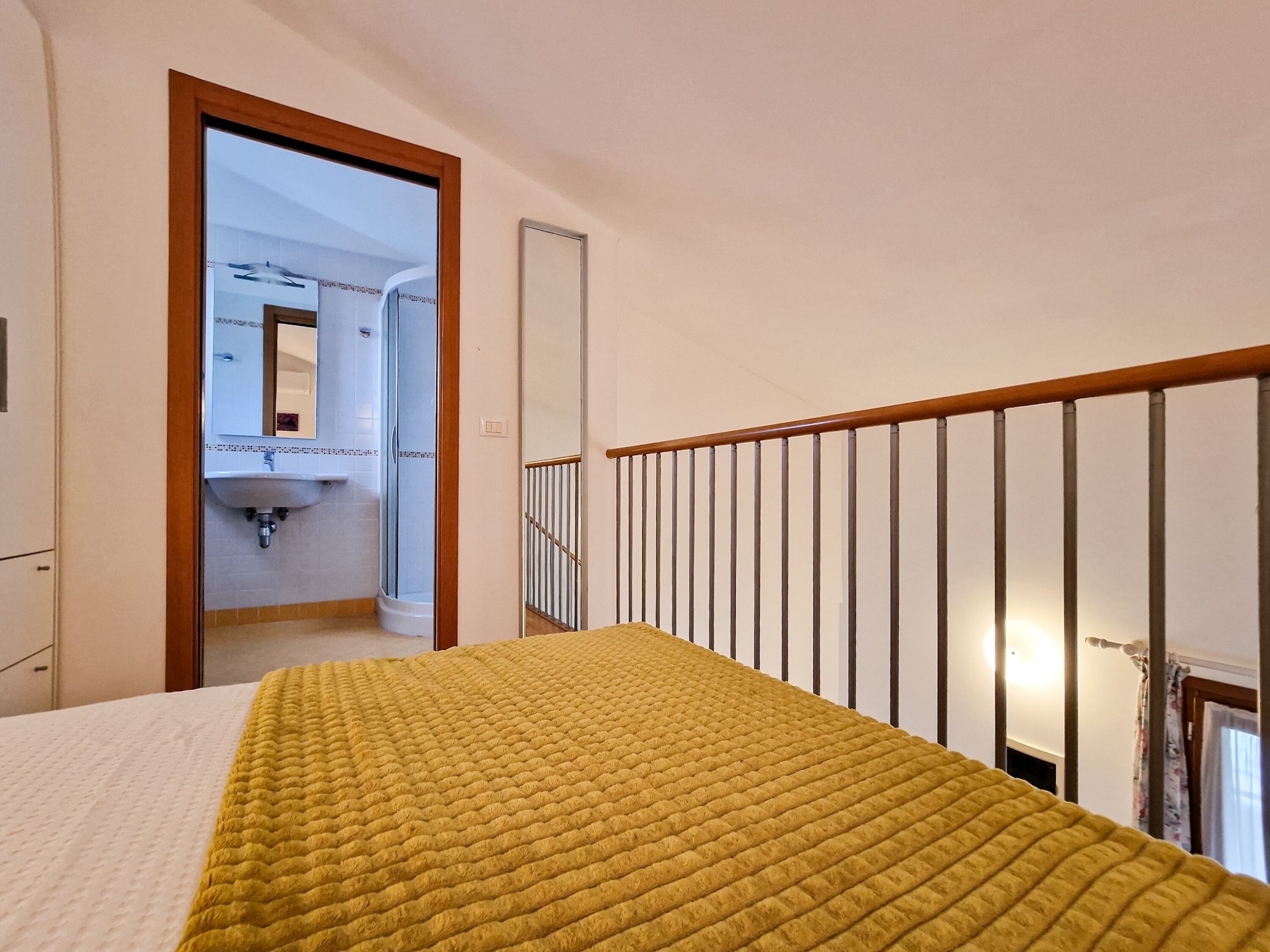Quarto de hotel com cama, cabeceira de madeira, banheiro com pia e espelho, e escada.