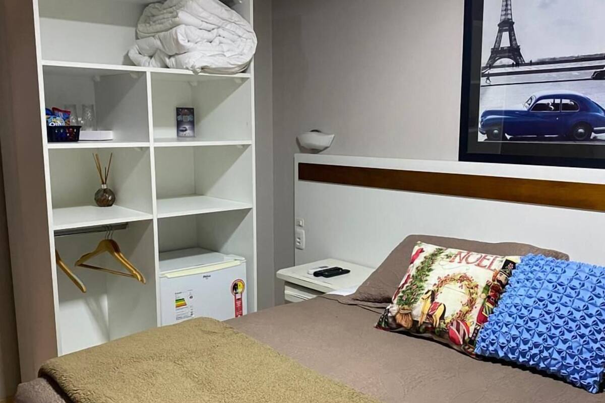 Ein Hotelzimmer mit Bett, Kleiderschrank, Schreibtisch, Stuhl, einem gerahmten Bild der Eiffelturms und einem Fenster.