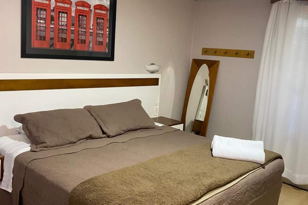 Ein Hotelzimmer mit einem Bett, einem gerahmten Bild roter Telefonzellen, einem Spiegel und einem Fenster mit Vorhängen.