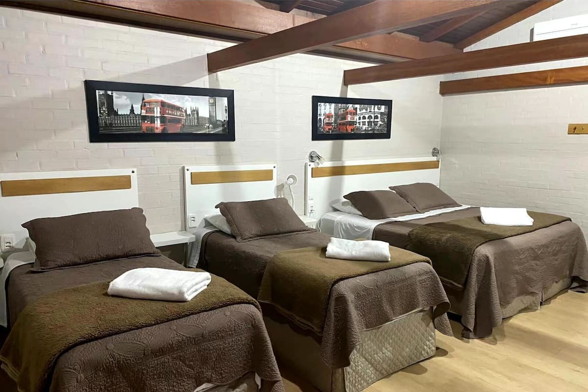 Ein Hotelzimmer mit drei Betten, jedes mit brauner Bettdecke und Kissen, und einem weißen Kopfteil. An der Wand hängen zwei Bilder in Rahmen: eines zeigt einen roten Doppeldeckerbus, das andere ein Gebäude mit Bögen. Das Zimmer verfügt über Holzbalken an der Decke und eine Wand aus weißen Ziegeln.