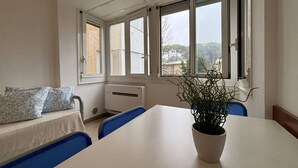 A room with a white table, blue chairs, a potted plant, a sofa with patterned cushions, and large windows with blinds. - Nice flat next to the beach in Bibione Pineda (Bibione)