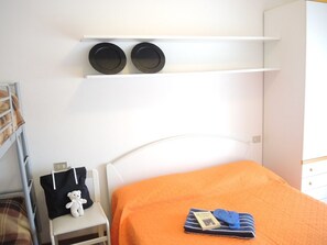 Une chambre à coucher avec un lit, une petite table, une étagère sur laquelle reposent deux objets noirs, et un ours en peluche sur un oreiller.