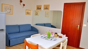 A compact living space with a blue sofa, a small dining table with a vase of flowers, and a red door. - Adorable flat with large terrace overlooking the sea (Bibione)