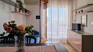 A modern apartment with a dining area, a living room, and a kitchen. There is a large window with sheer curtains, a flat-screen TV on the wall, and a potted plant on the shelf. - Homely apartment with great view and pool (Bibione)