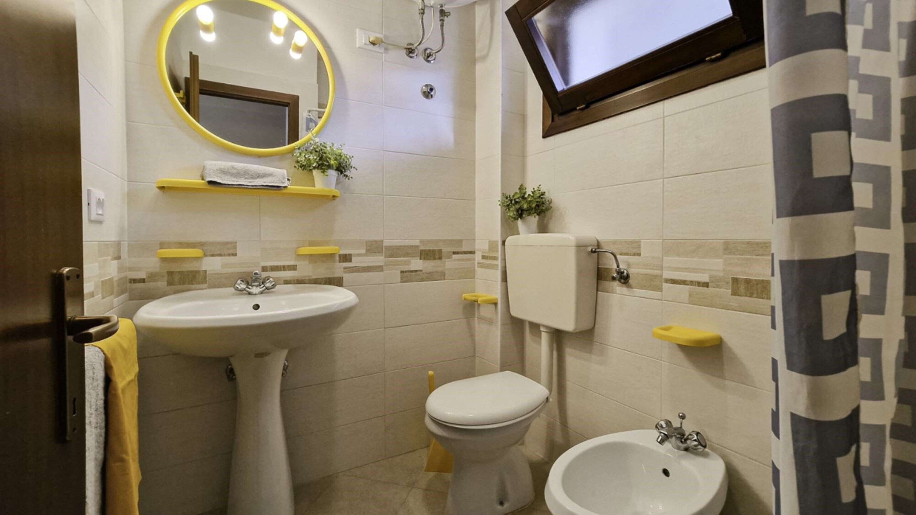 Een badkamer met een wastafel op een zuil, een toilet, een bidet, een spiegel met verlichting, en een douchegordijn.