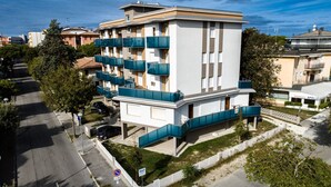Un immeuble résidentiel à plusieurs étages, doté de balcons, d’un petit espace jardin et d’une pizzeria appelée « Pizzeria Apollo », située à côté.