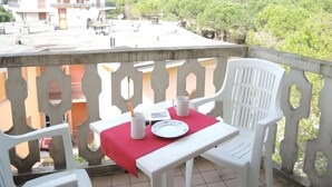 Un balcon avec une table dressée pour deux personnes, deux chaises en plastique blanches, une nappe rouge et deux tasses blanches.