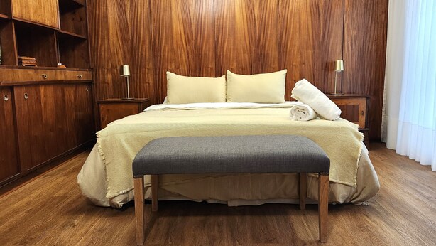 Een netjes ingerichte slaapkamer met een houten garderobe, een bed met een beige deken, een grijze bank en een witte handdoek ernaast.