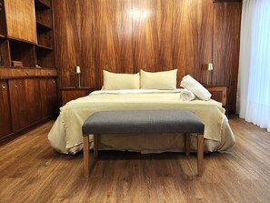 Een netjes ingerichte slaapkamer met een houten garderobe, een bed met een beige deken, een grijze bank en een witte handdoek ernaast.