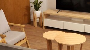 Een moderne woonkamer met een houten vloer, een grijze bank, twee ronde houten tafels, een televisie op een houten dressoir, en een potplant.