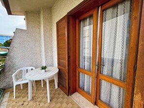 Uma varanda com uma cadeira e mesa de plástico branco, uma porta de madeira com painéis de vidro e uma parede texturizada.