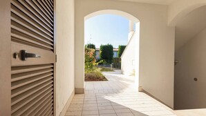 Un couloir doté d’un sol carrelé, une porte avec une poignée et un passage en arc donnant sur un espace extérieur avec un chemin et de la verdure.