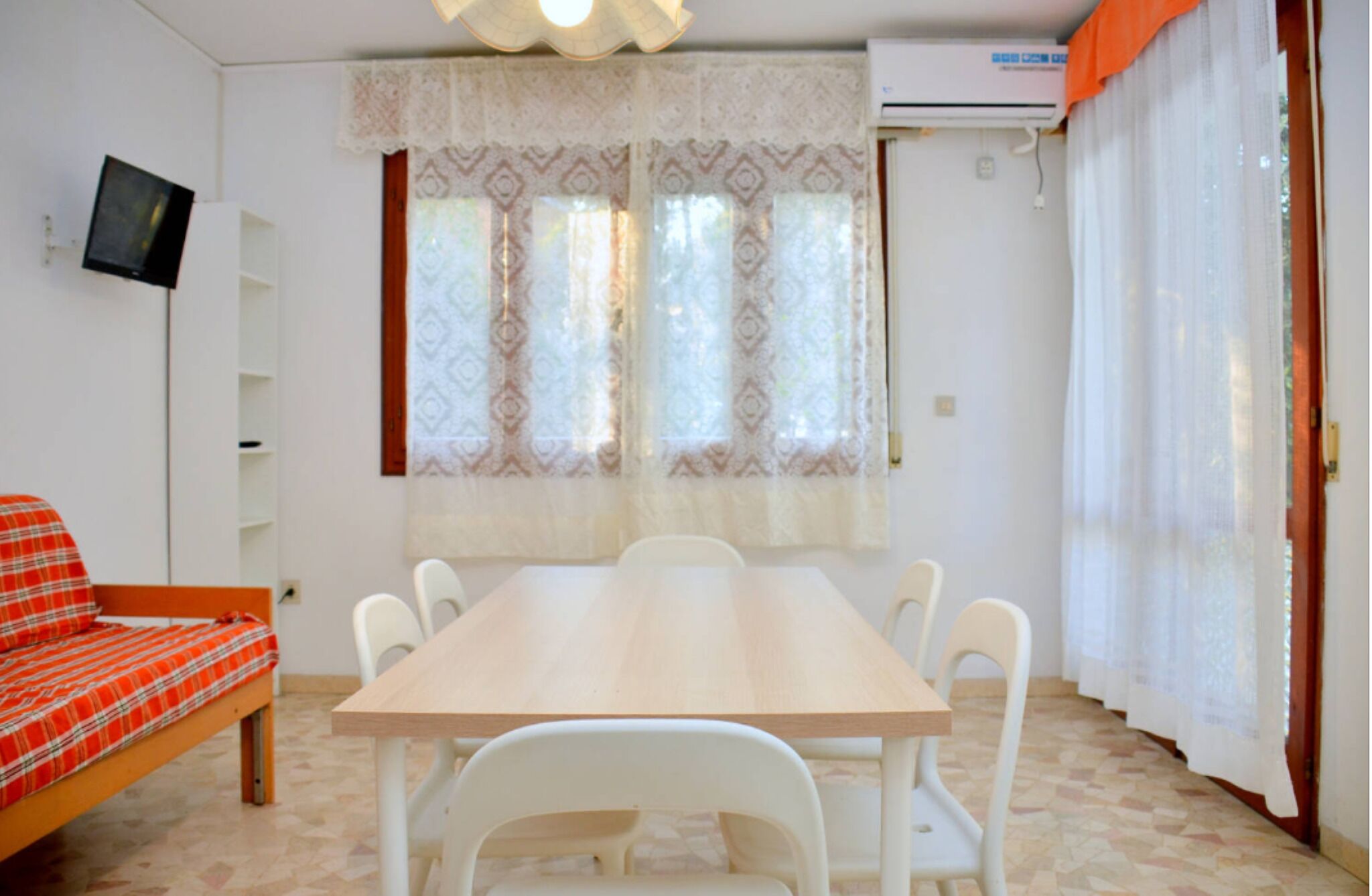 A room with a wooden table and white chairs, a red plaid sofa, and a television on the wall.