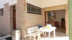 Un patio con una mesa y banco blancos, un refrigerador pequeño y una puerta corrediza de vidrio que da a una habitación interior.