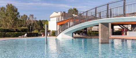 A swimming pool with a wooden deck in the foreground, a bridge with railings, and lounge chairs along the poolside.
