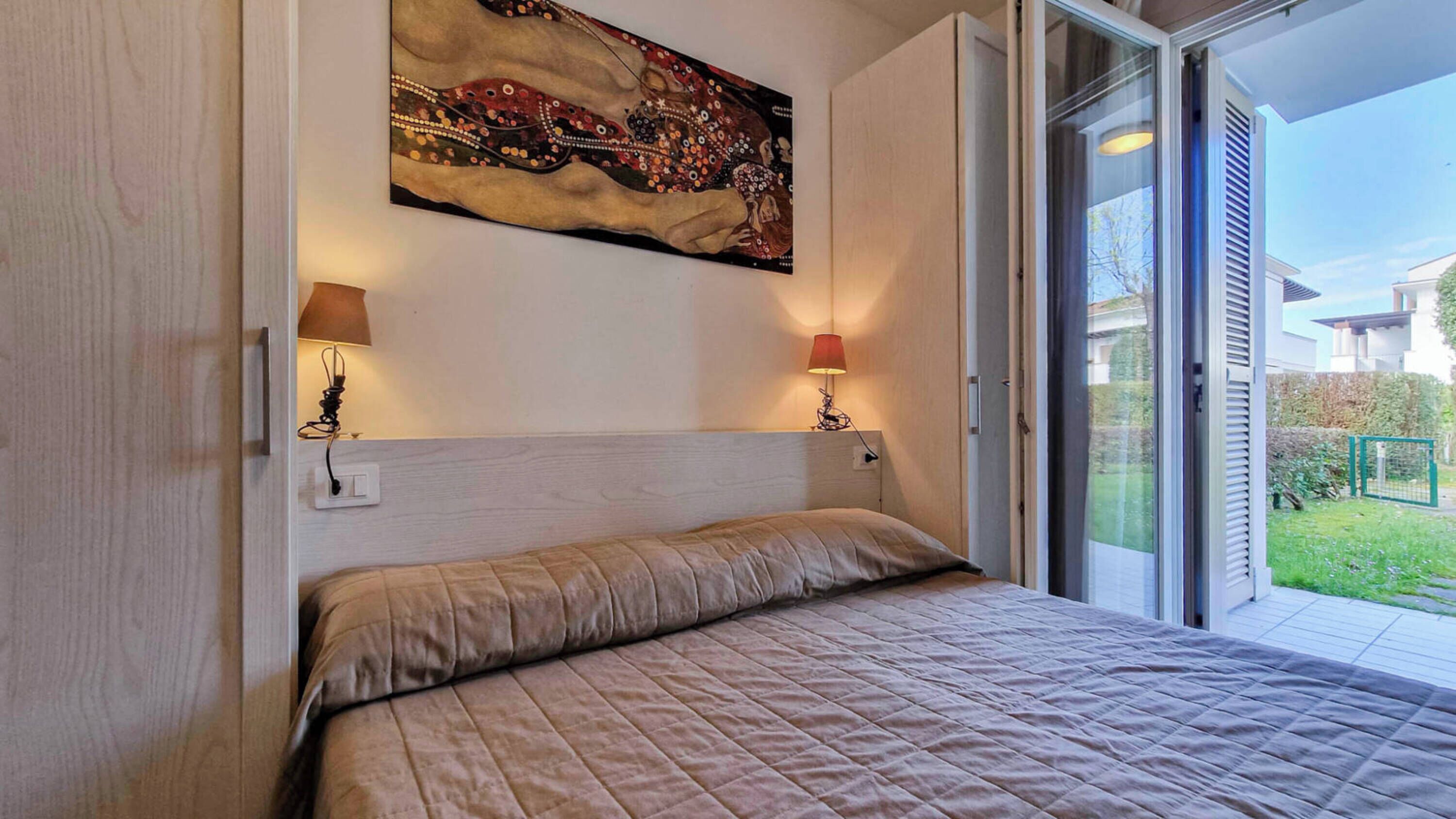 Een slaapkamer met een bed, twee bedlampen, een schilderij aan de muur en een glazen schuifdeur naar een balkon.