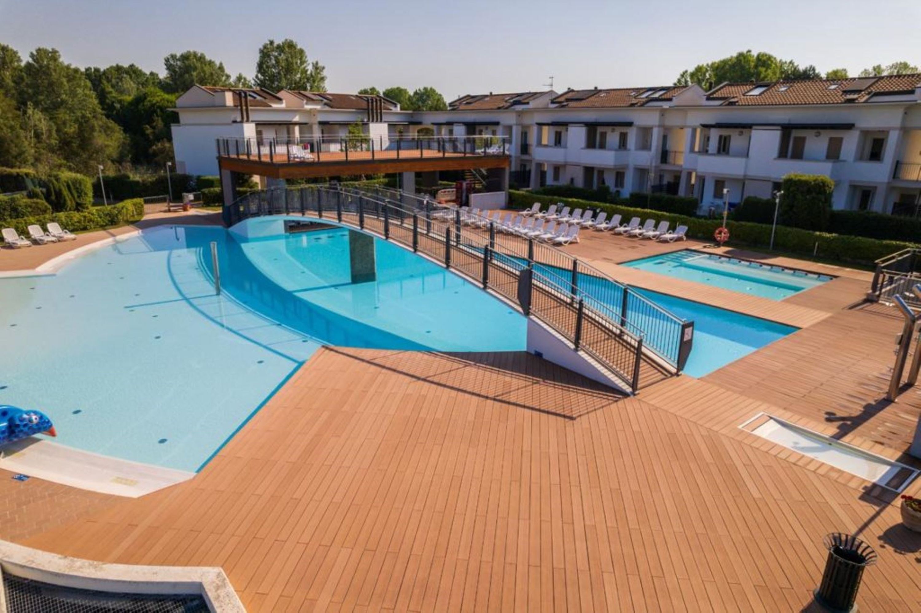 Une piscine entourée d’une terrasse en bois, de chaises longues et d’un bâtiment doté de plusieurs balcons.