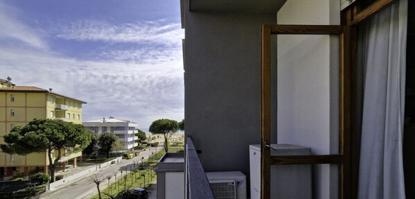En balkon med udsigt til en gade og bygninger, et vindue med gardin, og en aircondition.