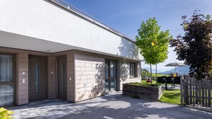 A modern building with a flat roof, large windows, and a paved entrance area with a bench and planter. - wohngenuss apprtements (Schladming)