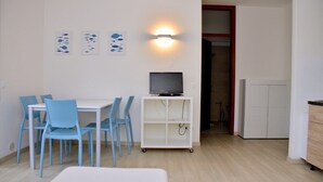 Ein kleiner, moderner Raum mit einem weißen Tisch und blauen Stühlen, einem weißen Schrank mit Fernseher und Kunstwerken an der Wand.