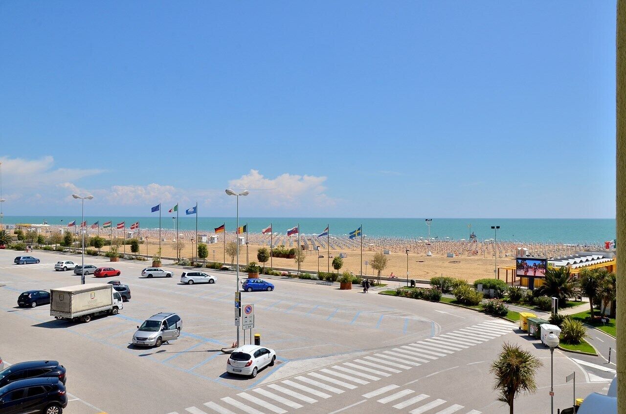 Ein Parkplatz mit Autos und einem Lastwagen, ein Strand mit Flaggen und ein klarer, blauer Himmel.