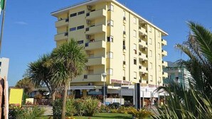 Un hôtel à plusieurs étages doté de balcons, un restaurant situé au rez-de-chaussée et un ciel d’un bleu éclatant.