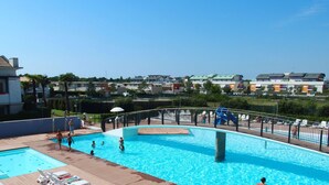 Ein Schwimmbecken mit Holzterrasse, umgeben von einem Metallzaun, mit Blick auf eine Wohngegend und verschiedenen Gebäuden.