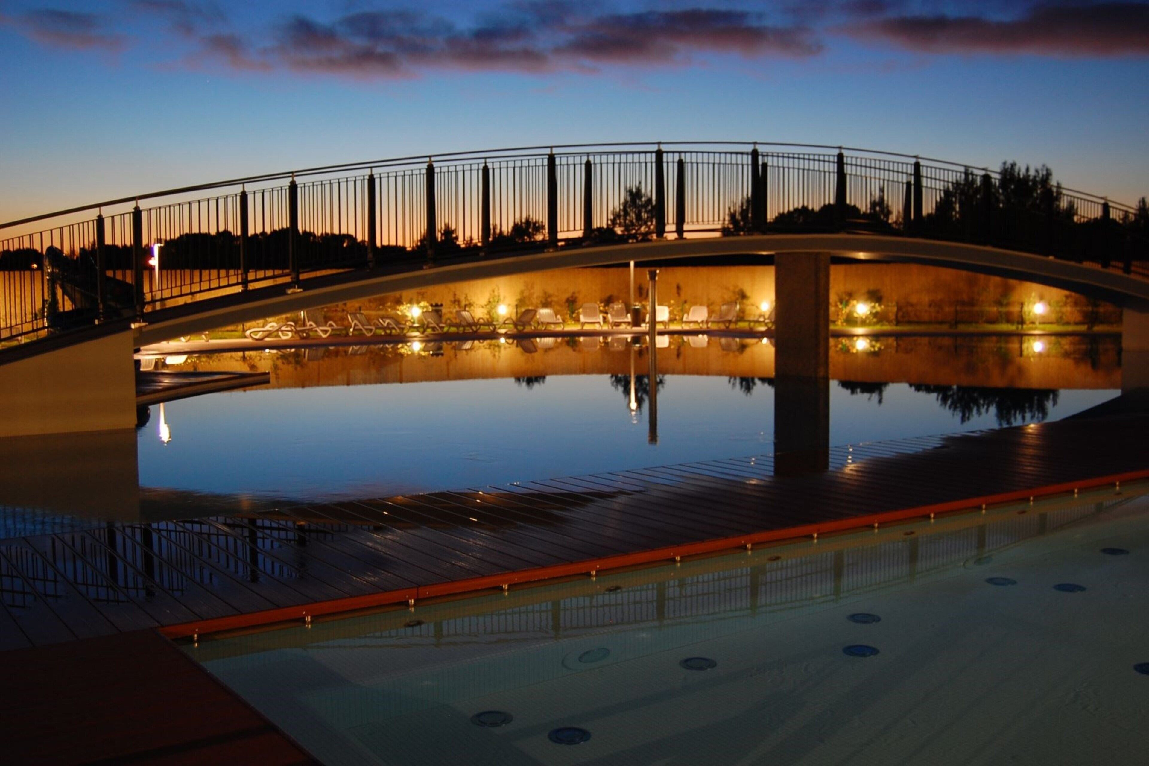 A bridge over a reflective pool with a railing and lights, surrounded by trees.