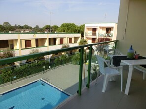Un balcon avec une piscine, des chaises en plastique blanches et une table sur laquelle se trouvent une bouteille et une canette.