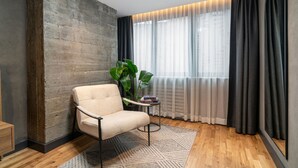 A modern room with a concrete accent wall, a beige armchair, a small side table with books, a potted plant, and a large window with sheer curtains. - Urban Quarters - Gülhane (Istanbul)
